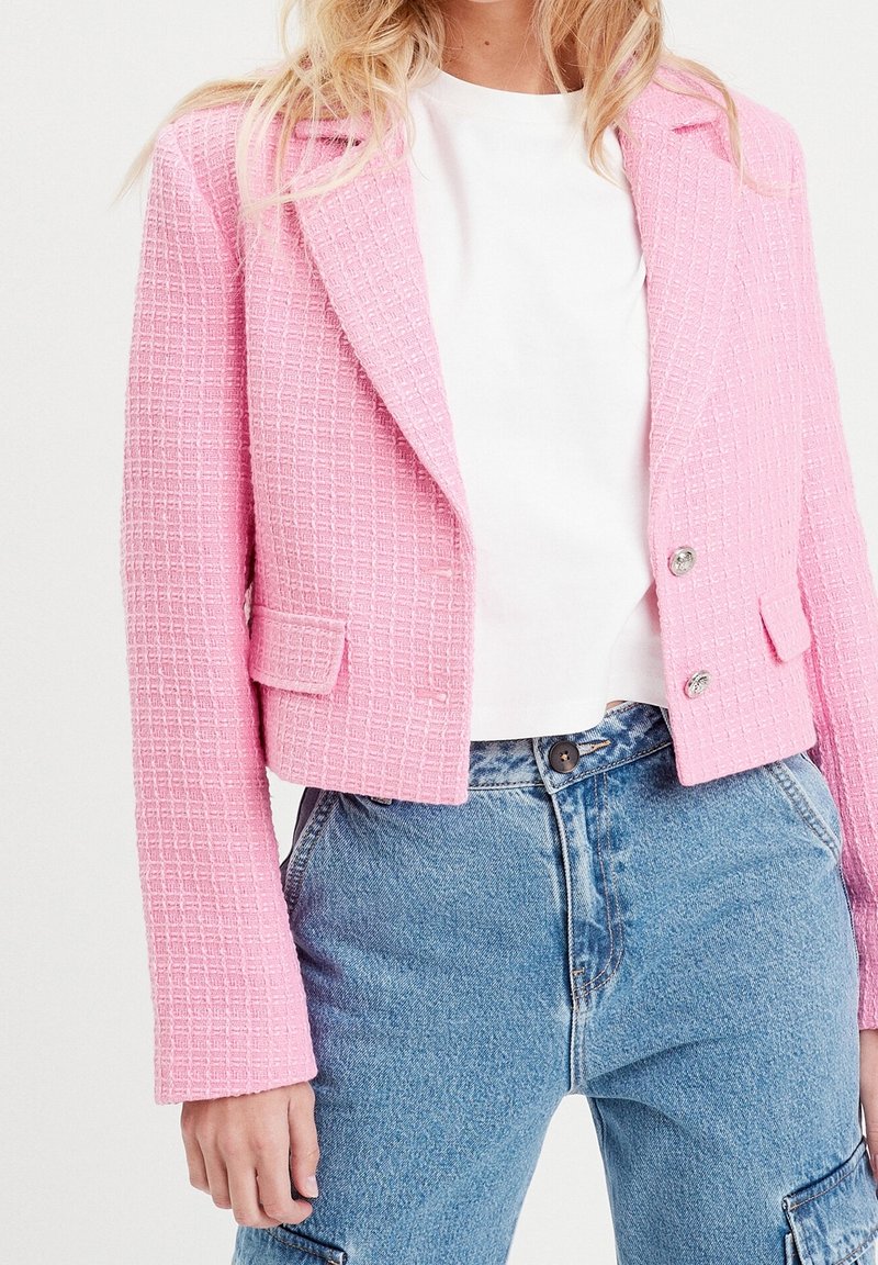 Blazer en tweed rose à surface texturée, avec des revers en cranté, deux poches avant et des boutons argentés, porté avec un t-shirt blanc et un jean bleu.