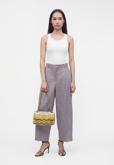 Femme debout portant un haut blanc sans manches, un pantalon lavande à jambes larges, des talons noirs à lanières, tenant un sac à main jaune à motif zigzag.