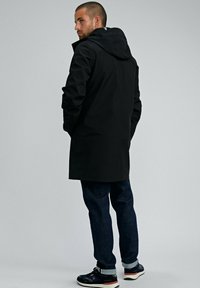 Homme en manteau à capuche noir, jeans foncés et baskets, se tenant les mains dans les poches, regardant par-dessus son épaule contre un fond uni.