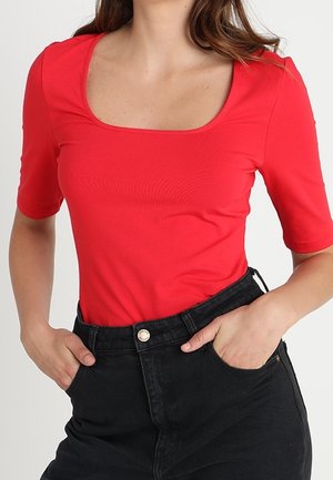 Vrouw draagt een nauwsluitende felrode top met een vierkante hals en ellebooglange mouwen, gecombineerd met een zwarte high-waist jeans, handen in de zakken.