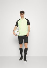 Camiseta de fútbol de manga corta en verde y negro, con pantalones cortos y calcetas negras hasta la rodilla, sosteniendo un balón de fútbol con un patrón, contra un fondo liso.