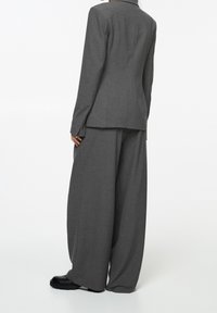 Veste de costume grise sur mesure à manches longues et silhouette ajustée, associée à un pantalon gris à jambes larges et des chaussures noires.