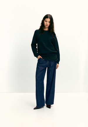 Sort sweater med løs pasform, kombineret med vidde ben i mørk denim. Modellen står med den ene hånd i lommen, hvilket fremhæver stoffets tekstur og farvekontrast.