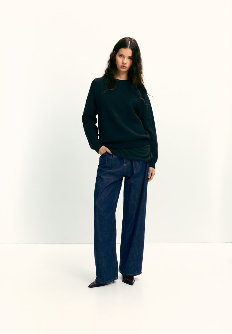 Fekete pulóver laza szabással, széles szárú sötét farmernadrággal párosítva. A modell az egyik kezét a zsebében tartja, kiemelve a szövet textúráját és a színkontrasztot.