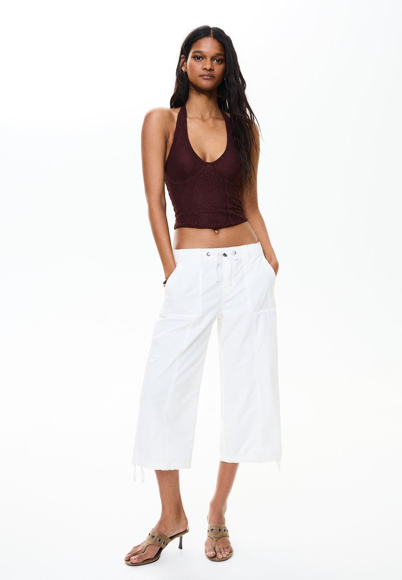 Woman stands with hands in pockets wearing sleeveless brown crop top, white wide-leg pants, and open-toe heeled sandals on white background.