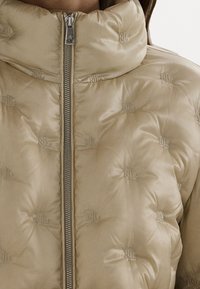 Kurtka puchowa w jasnobeżowym kolorze z błyszczącym wykończeniem, ozdobiona haftowanymi wzorami logo, z wysokim kołnierzem i centralnym zamkiem błyskawicznym.