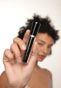 Tube cosmétique noir avec base transparente, accent argenté, tenu par une main avec un vernis à ongles clair et naturel. Étiquetage clair sur le produit.