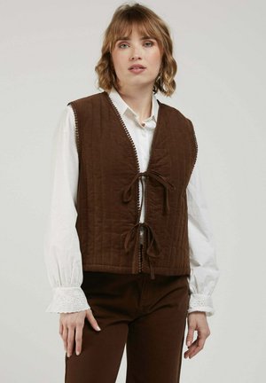 Femme portant un chemisier blanc à manches longues avec des poignets en dentelle sous un gilet matelassé marron noué à l'avant, associé à un pantalon marron.