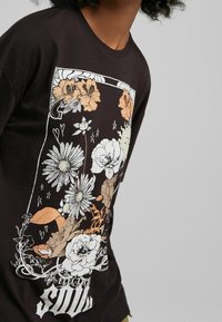 Mustat T-paidat, joissa on kukkakuosisuunnittelu valkoisena, oranssina ja vihreänä. Pyöreä kaula-aukko ja rento istuvuus, pehmeä kangas.