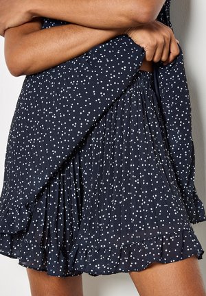 Hände, die einen marineblauen, gepunkteten, plissierten Rock mit einem gerafften Saum an einer Person vor einem schlichten weißen Hintergrund justieren.