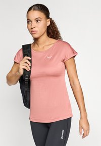 Īsām piedurknēm ar apaļu kakla izgriezumu, izgatavota no mīksta rozā auduma ar mazu balto logotipu; valkāta kopā ar melniem sporta biksēm un melnu mugursomu.