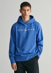 Felpa blu in cotone con tasca a canguro, con il logo "GANT 1949" in bianco e un design a scudo. Tessuto morbido e vestibilità rilassata.