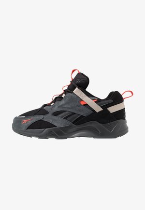 Scarpa da ginnastica nera e grigia con dettagli arancioni, pannelli in mesh e suede, cinturino beige sul mesopiede e suola spessa e testurizzata.