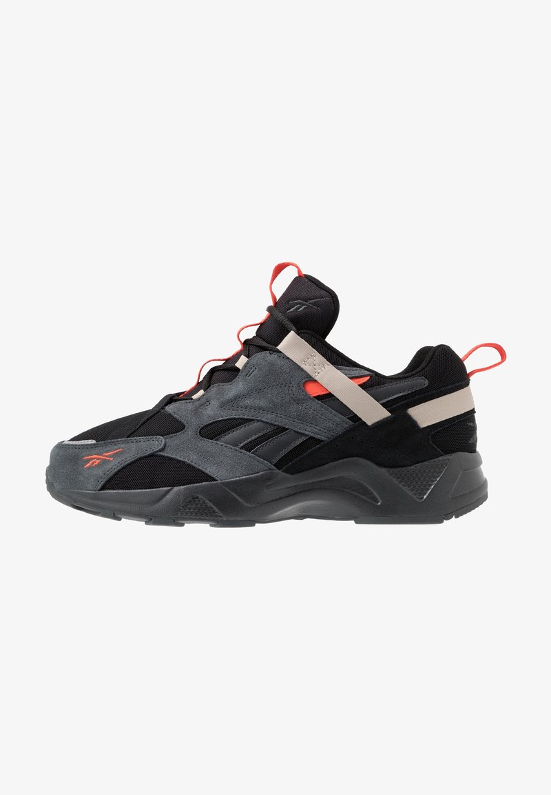 Baskets athlétiques noires et grises avec des accents orange, panneaux en mesh et en daim, une lanière beige sur le médio-pied, et une semelle épaisse texturée.