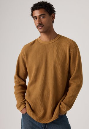 Homme aux cheveux bouclés et moustachu, portant un pull en tricot marron et un jean bleu, debout les mains dans les poches sur un fond uni.