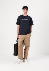 Tengerészkék pamut póló "BELSTAFF" felirattal, világosbarna nadrággal és fehér sportcipőkkel párosítva. Fekete vászontáskát tart. Egyszerű dizájn.