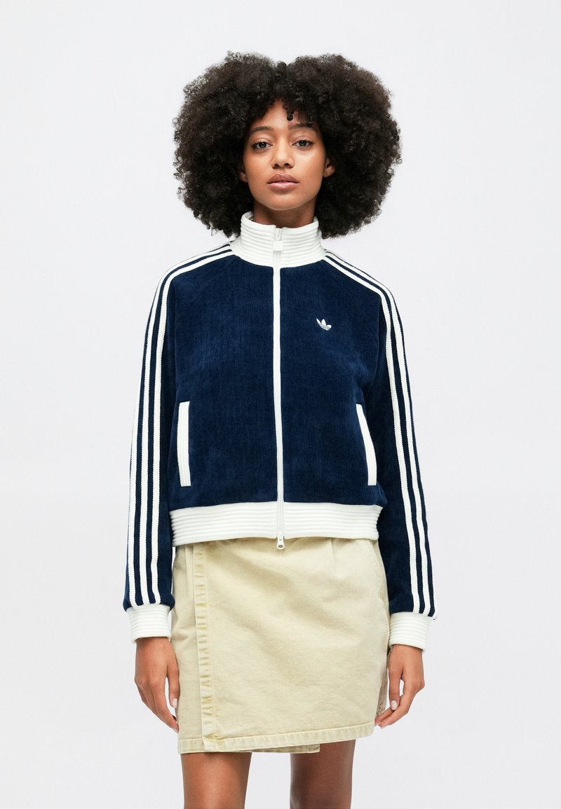 Veste en velours côtelé bleu marine avec un col haut, une fermeture éclair blanche et des rayures. Dispose de poches zippées blanches et d'un logo brodé sur la poitrine.