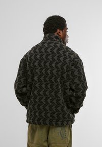 Giacca in pile nera e grigia con un alto colletto e un design a zigzag testurizzato, indossata sopra pantaloni verde oliva.