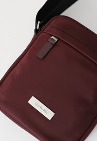Burgundy crossbody taske lavet af slidstærkt stof; har et elegant design, metalhardware og et synligt Calvin Klein logo på en sølvfarvet detalje.
