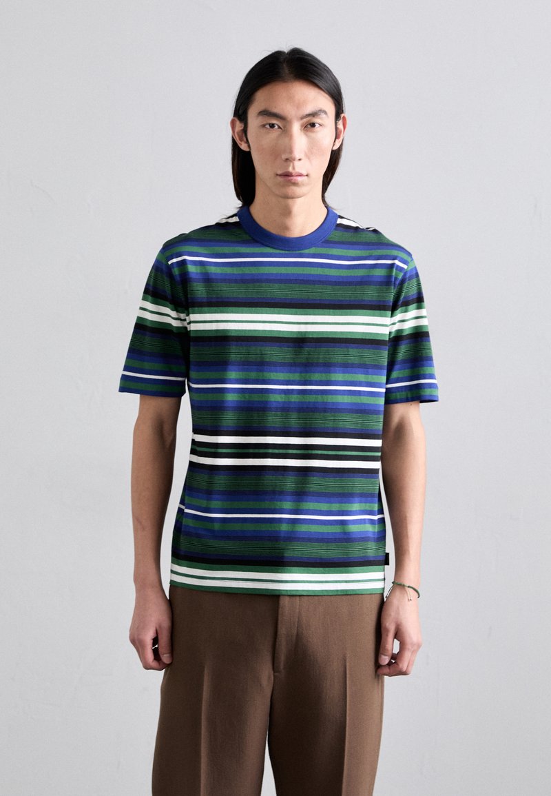 T-shirt à rayures à manches courtes en bleu, vert, noir et blanc. Doté d'un col rond et d'un tissu texturé. Porté avec un pantalon marron.