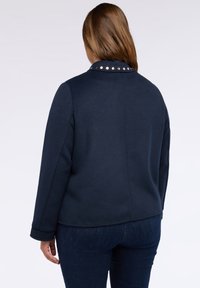 Femeie cu păr lung, brunet, îmbrăcată într-o jachetă albastră închisă cu studs metalici pe guler, întoarsă cu spatele, pe un fundal deschis simplu.