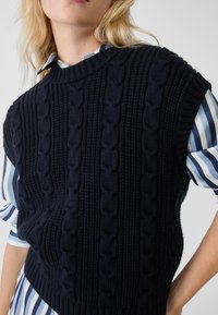 Blond kvinna som bär en marinblå kabelstickad ärmlös tröja över en blå och vit randig skjorta, med handen på midjan.