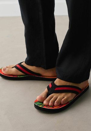 Füße in schwarzen und roten Flip-Flops mit bunten Sohlen, kombiniert mit schwarzen Hosen, stehend auf einem hellgrauen Boden.