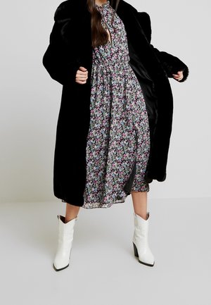 Kvinde iført en sort faux pelsfrakke over en blomsterprint midi kjole og hvide cowboystøvler med hæl, stående mod en ensfarvet baggrund.