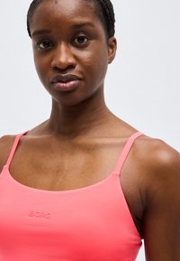 Νέα γυναίκα με πλεξούδες, που φοράει τοπ κοραλλί ροζ με λεπτές τιράντες, σε απλό φωτεινό φόντο.