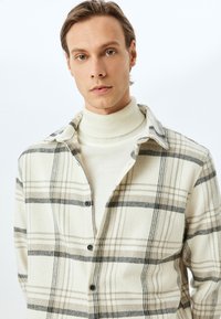 Beige geruite overhemd met zwarte en bruine lijnen; voorzien van een kraag en zwarte knopen, gedragen over een crèmekleurige coltrui.