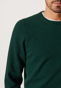 Mörkgrön stickad tröja med rund halsringning, slät yta och åtsittande ärmar, visad mot en ljus bakgrund.