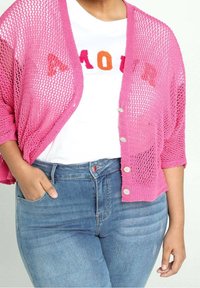 Lätt, öppet stickad rosa kofta med trekvartslånga ärmar och knappknäppning, bärs över en vit T-shirt med färgglad text, ihop med blå jeans.