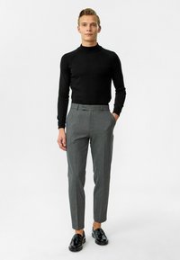 Zwarte aansluitende coltrui, grijze op maat gemaakte broek en zwarte glanzende instappers. Soepele stof met een elegant ontwerp. Neutrale achtergrond.