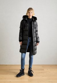 Giacca a vento nera con cappuccio, caratterizzata da un esterno lucido e elegante, polsini a coste e una lunghezza lunga, abbinata a jeans blu e sneakers alte nere.