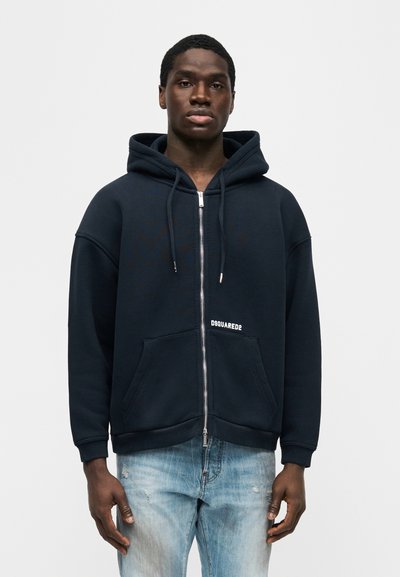 Νεαρός άνδρας που φοράει μπλε σκούρο μπουφάν με φερμουάρ και το λογότυπο "DSQUARED2" και ανοιχτόχρωμα τζιν με φθορές, στέκεται μπροστά σε απλό φόντο.