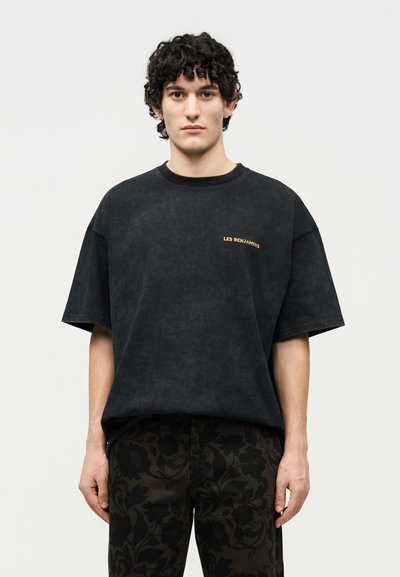 Νεαρός άνδρας με σγουρά μαλλιά, φορώντας μια άνετη μαύρη μπλούζα με το κείμενο "LES BENJAMINS" και σκούρο παντελόνι με λουλουδάτο σχέδιο, στέκεται μπροστά σε ένα απλό φόντο.