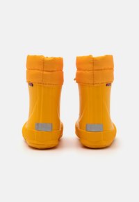 Bottes en caoutchouc jaunes avec un revers en tissu orange rembourré, motif circulaire sur la semelle et accents argentés au talon. Vue arrière.