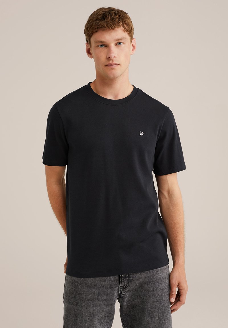 Jaunas vyras su trumpais garbanotais plaukais, vilkintis paprastą juodą marškinėlį su mažu paukščio logotipu kairėje krūtinės pusėje ir pilkomis džinsais, stovi priešais kreminę foną.