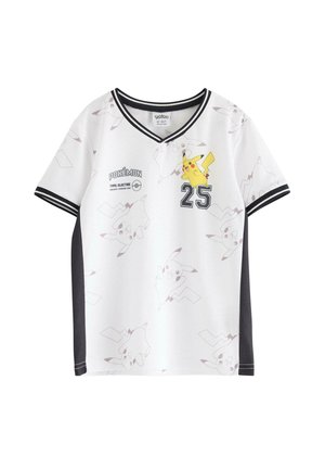 Ein kleines Kind steht lächelnd da, trägt ein weißes Pikachu-Shirt mit schwarzem Rand, schwarze Hosen mit weißen Streifen und beigefarbene Sneakers.