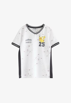 Ein kleines Kind steht lächelnd da, trägt ein weißes Pikachu-Shirt mit schwarzem Rand, schwarze Hosen mit weißen Streifen und beigefarbene Sneakers.