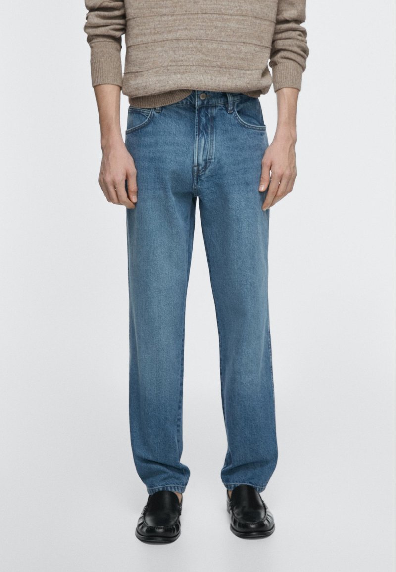 Personne portant un jean bleu ample, un pull beige et des chaussures en cuir noires, debout devant un fond gris clair uni.