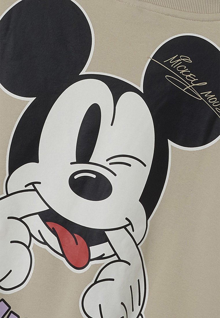 T-shirt beige con una grande stampa grafica di un personaggio dei cartoni animati con orecchie nere, viso bianco e un'espressione giocosa, mentre fa le linguacce.