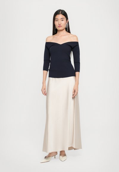 Tumes off-the-shoulder ribased top kolmekvartaliste varrukatega, mis on ühendatud sileda kreemikarva maksikleidiga ja valgete terava ninaga kingadega.