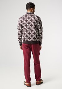 Mönstrad stickad tröja i mörkbrunt med vita och mörkröda lövmotiv, i kombination med mörkröda byxor och randiga bruna och orange skor.