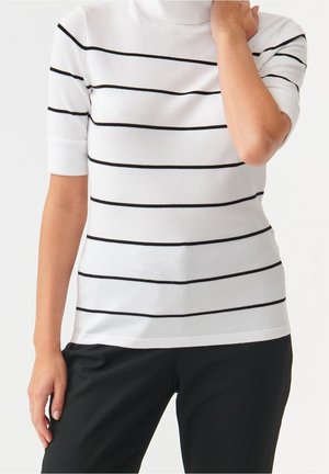 Nainen valkoisessa lyhythihaisessa pooloneuleessa, jossa mustat vaakaraitarit, ja mustissa housuissa, käsi koskettaa kaulaa.