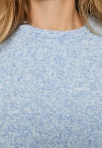 Gaiši zils džemperis ar mīkstu, teksturētu adījumu un apaļu kakla izgriezumu, kuram visā virsmā ir smalka pleķaina raksts.