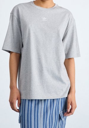 Persoon die een lichtgrijs Adidas T-shirt met korte mouwen en een blauw-witte verticale gestreepte broek met zakken draagt.