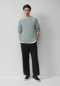 Hellsgrüner Strickpullover mit Raglanärmeln, kombiniert mit dunkel lockerer Hose. Schwarze Sneaker und ein weißes Unterhemd sichtbar.