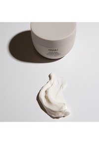 Romige witte haarsproduct in een ronde, lichtbeige container met een gladde deksel en tekst aan de zijkant; een klodder crème ernaast geplaatst.
