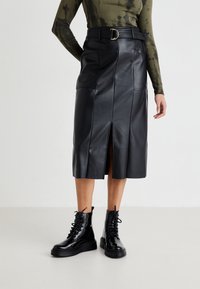 Jupe midi en cuir noir avec fente à l'avant, poches latérales et une large ceinture avec une boucle en or. Associée à des bottes de combat en dentelle noires.